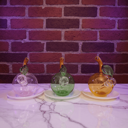 🍎 Apple Glass Bong – Mini Fruit Water PipeThree colorful glass bongs with leaf designs on a marble surface against a brick wall.