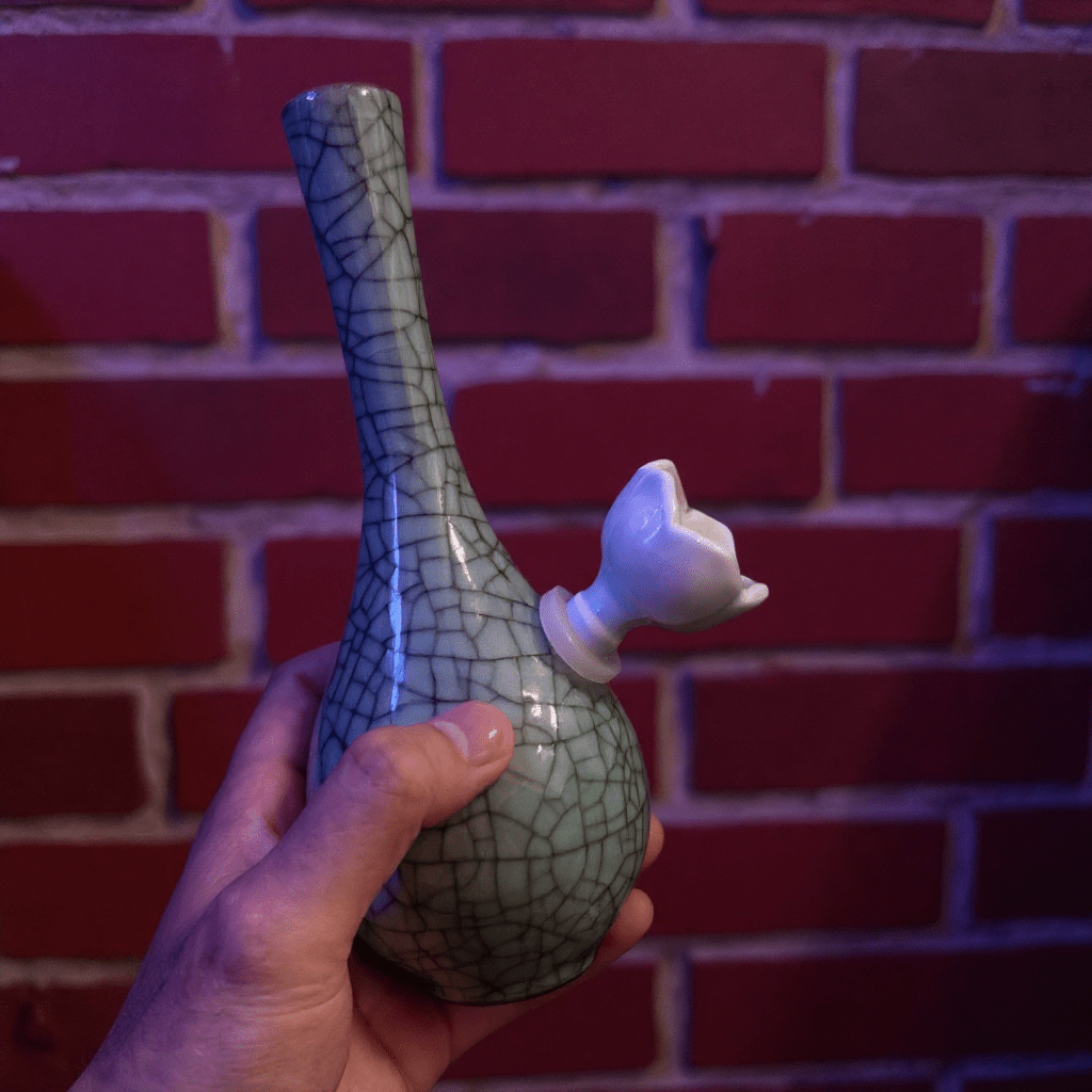 Crackle Glaze Slim Neck Celadon Porcelain BongHand holding a ceramic bong with a cracked glaze design against a brick wall.