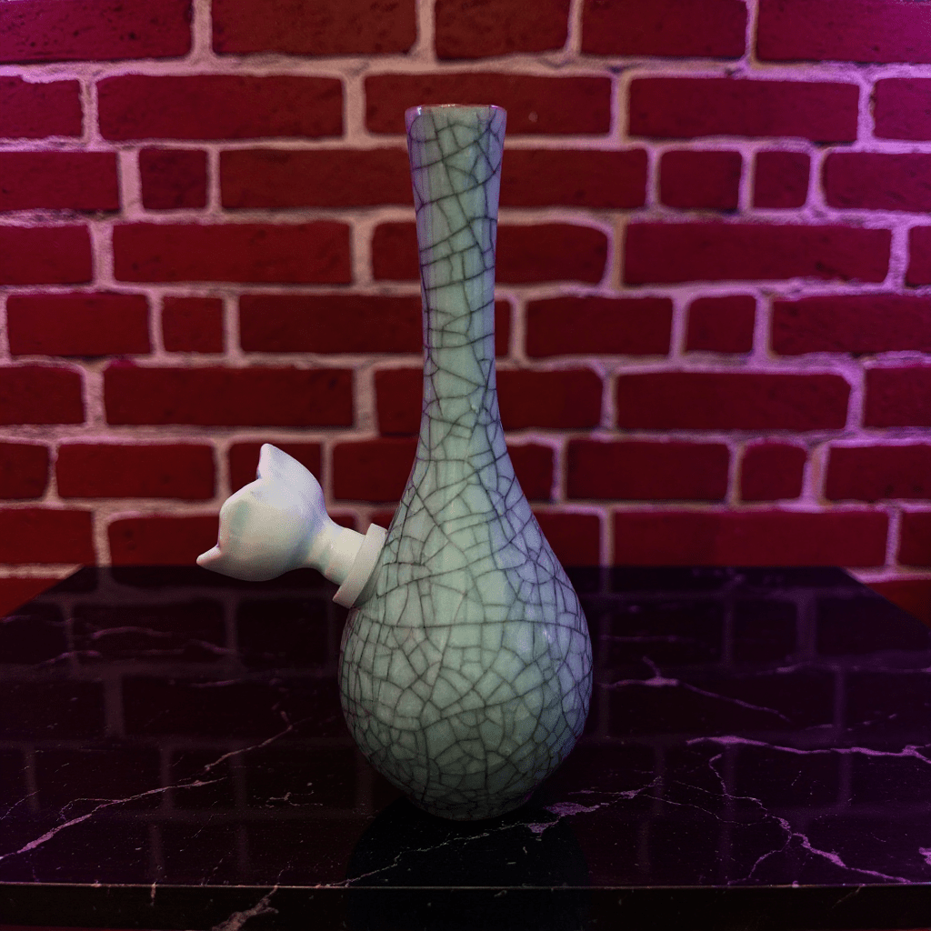 Crackle Glaze Slim Neck Celadon Porcelain BongGreen ceramic bong with textured surface on a dark surface against a brick wall background