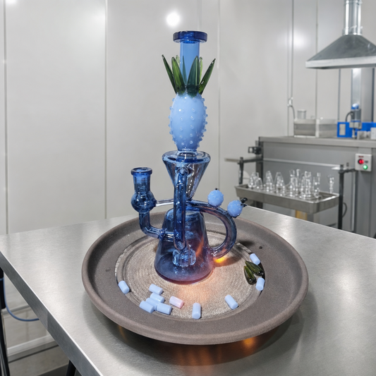 A blue glass water pipe with a pineapple-shaped top section and green leaf details, displayed on a round tray in a clean laboratory or workshop setting.