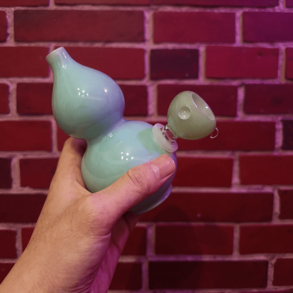 Glossy Blue Celadon Gourd Porcelain BongHand holding a light blue ceramic gourd - shaped bong against a red brick wall.