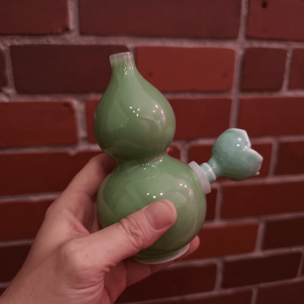 Glossy Green Celadon Gourd Porcelain BongGreen ceramic gourd - shaped bong held in front of a brick wall