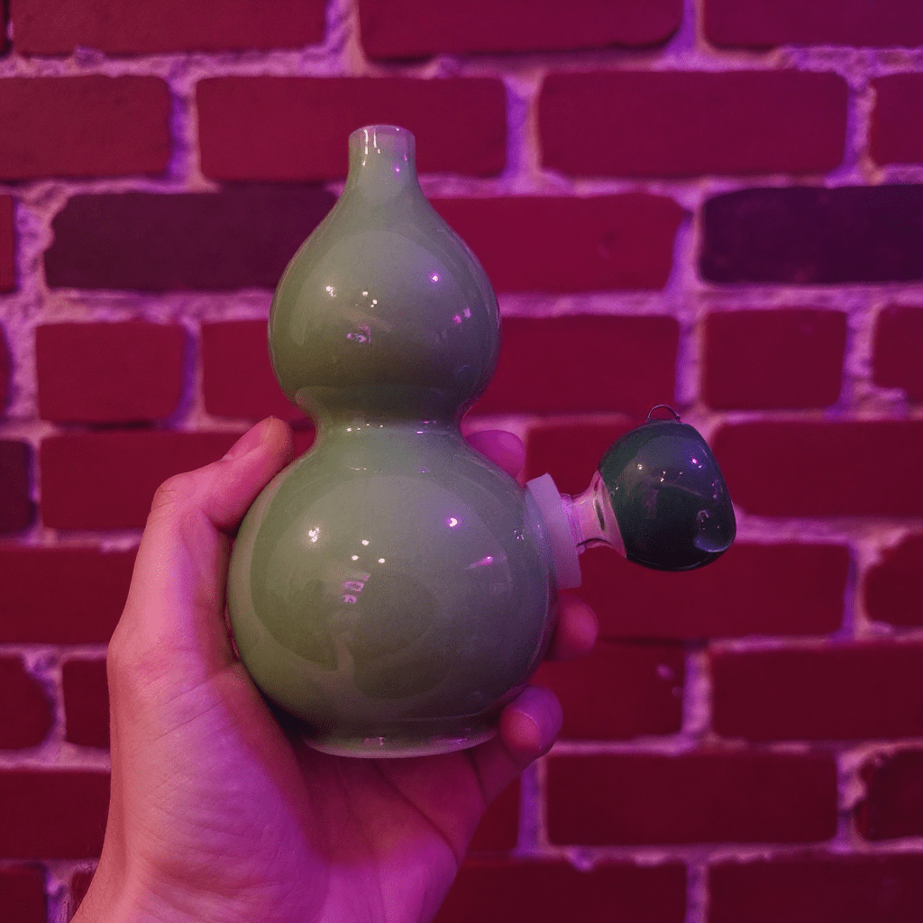 Glossy Green Celadon Gourd Porcelain BongGreen ceramic gourd - shaped bong held against a brick wall with purple lighting