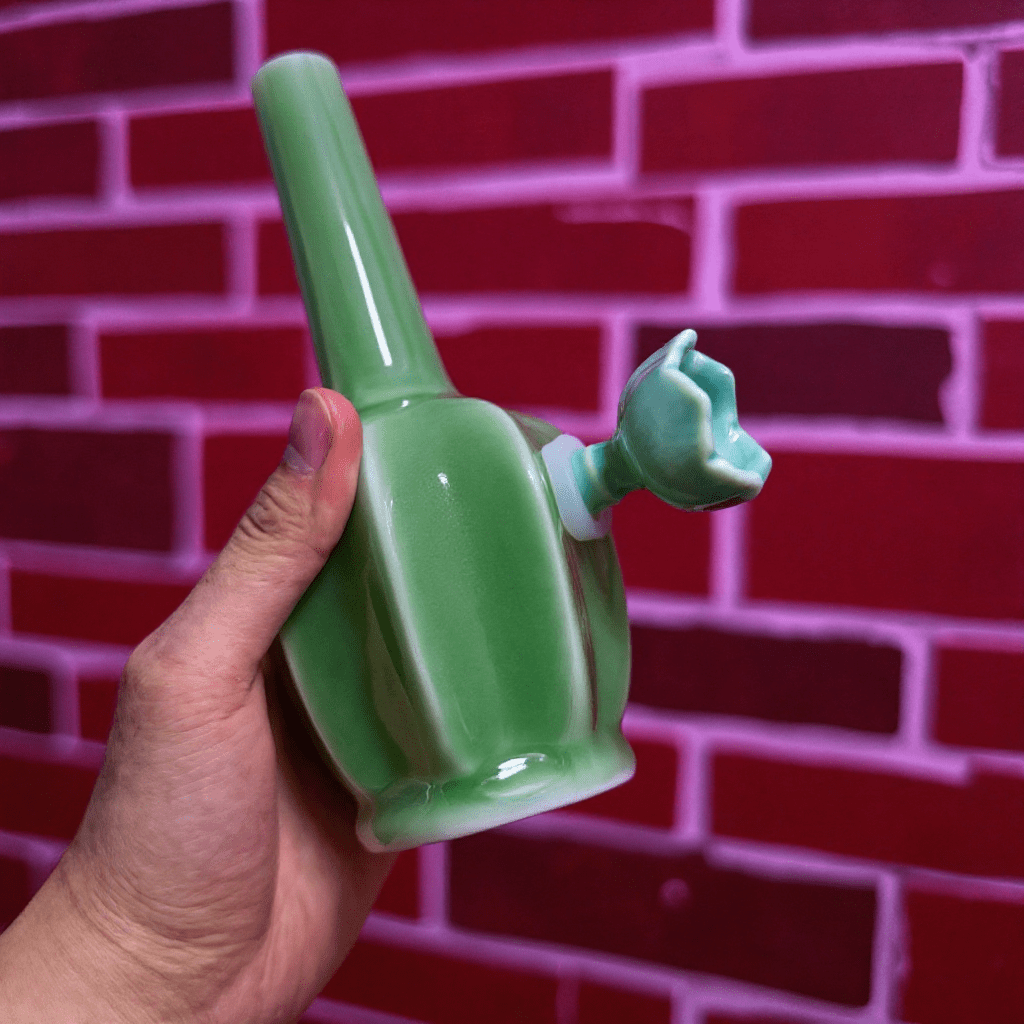 Glossy Green Flat - Sided Celadon Porcelain BongGreen ceramic pipe held by a hand against a red brick wall.