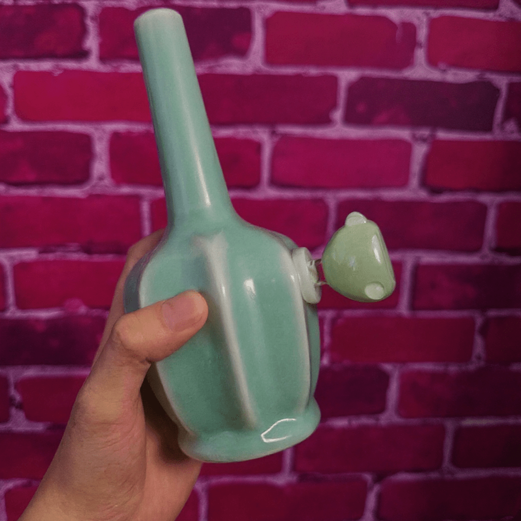 Glossy Light Blue Celadon Flat - Sided Porcelain BongGreen ceramic pipe held by a hand against a red brick wall.