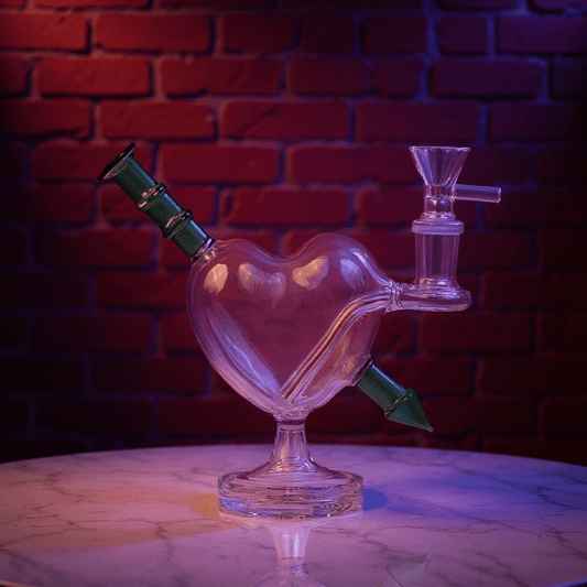 Heartpiercer Water Pipe – Mint Arrow EditionHeart - shaped glass smoking device with green accents on a reflective surface against a brick wall.