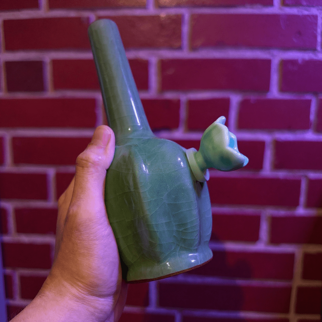 Ice - Crack Green Celadon Flat - Sided Porcelain BongGreen ceramic bong with ceramic bowl held by a hand against a brick wall.