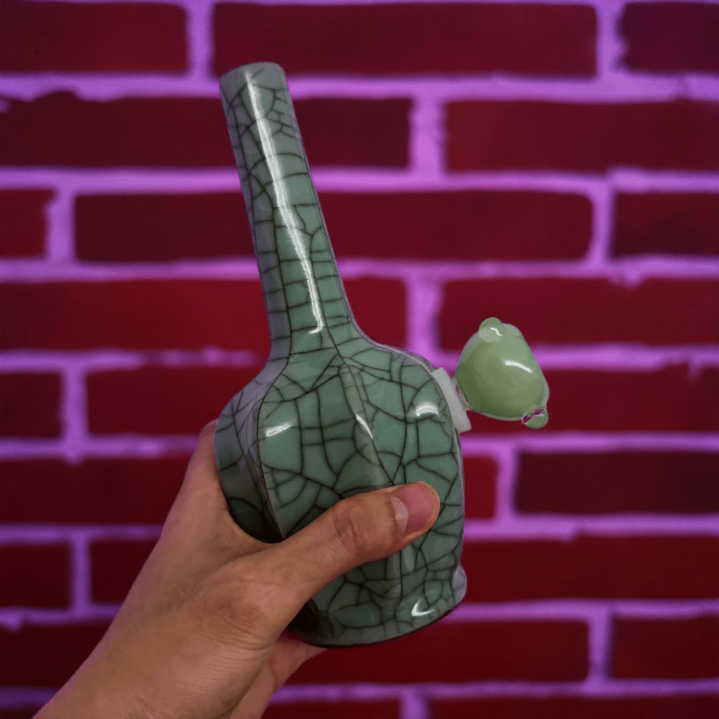 Ice - Crack Light Blue Celadon Flat - Sided Porcelain BongGreen ceramic bong with a unique texture held against a red brick wall.