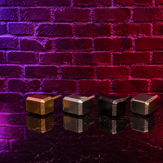 Thor Hammer Shape Grinder – 4-Part Metal GrinderFour miniature Thor's hammer (Mjolnfr) figurines in gold, silver, and black finishes lined up on a reflective dark surface against a purple and red brick wall background.