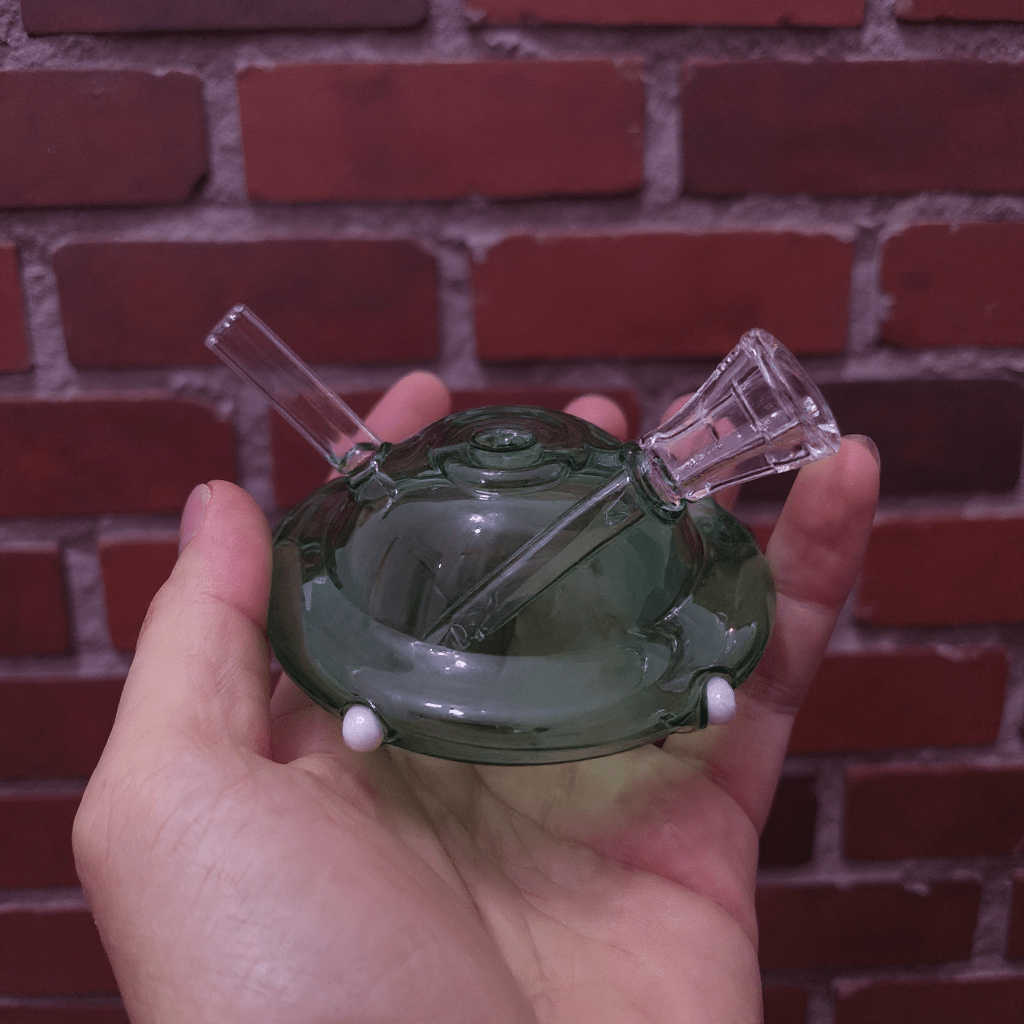 UFO Mini Glass Bong – Water Pipe in 3 ColorsHand holding a green glass smoking device against a brick wall background
