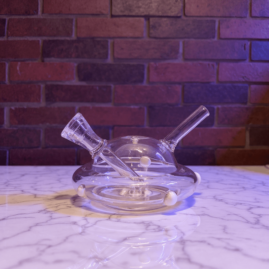 UFO Mini Glass Bong – Water Pipe in 3 Colorspink glass smoking bong on a marble surface with a brick wall background