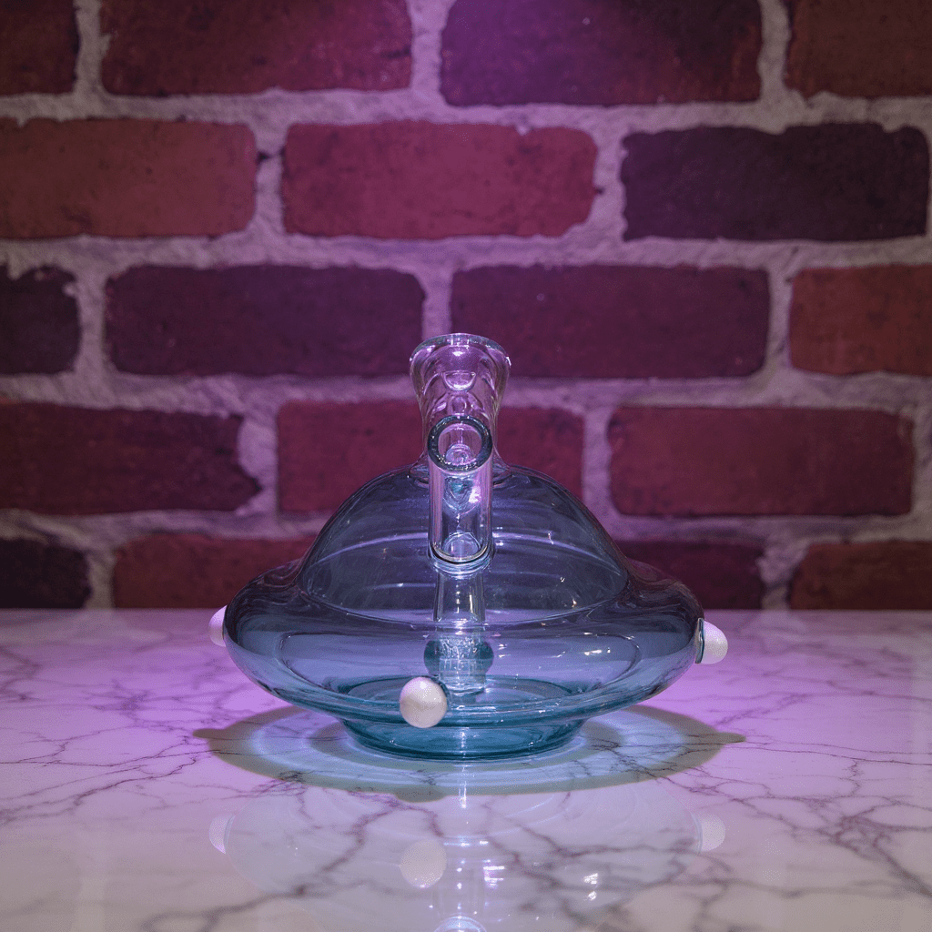 UFO Mini Glass Bong – Water Pipe in 3 Colorsblue glass object on a marble surface with a brick wall background