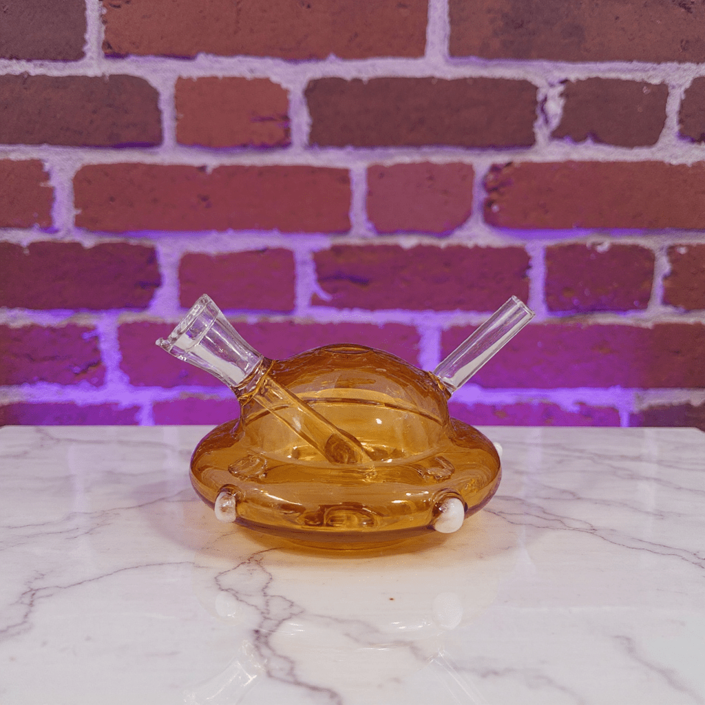 UFO Mini Glass Bong – Water Pipe in 3 ColorsAmber glass pipe with two clear hoses on a marble surface against a brick wall.