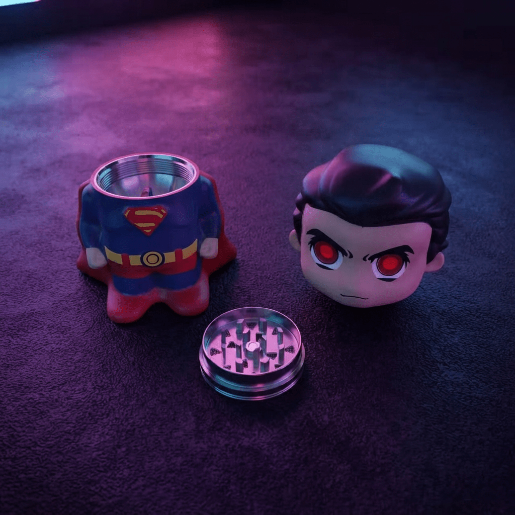 Caped Comic Hero Character Herb Grinder – Novelty Metal GrinderA Superman-themed grinder set on a dark textured surface under purple and pink lighting. The set includes a grinder shaped like Superman's chest/torso, a character head with red eyes, and a clear metal grinding chamber.
