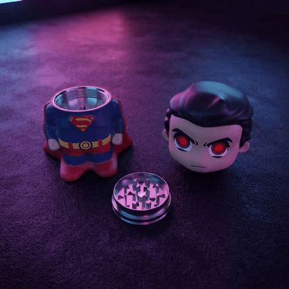 Caped Comic Hero Character Herb Grinder – Novelty Metal GrinderA Superman-themed grinder set on a dark textured surface under purple and pink lighting. The set includes a grinder shaped like Superman's chest/torso, a character head with red eyes, and a clear metal grinding chamber.