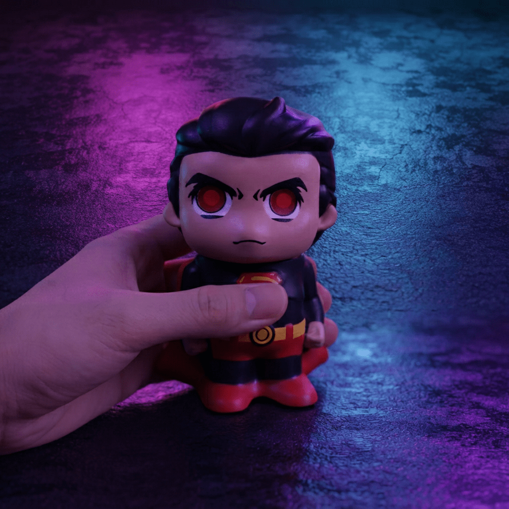 Caped Comic Hero Character Herb Grinder – Novelty Metal GrinderA hand holding a small superhero-style vinyl figure against a dark textured background with purple and blue lighting. The figure has dark hair, red eyes, a black top, and red and yellow accents.