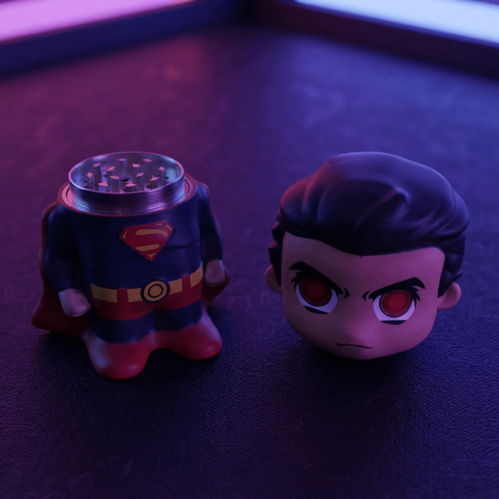 Caped Comic Hero Character Herb Grinder – Novelty Metal GrinderA Superman-themed smoking accessory set featuring a grinder shaped like Superman's chest and a small Superman character head figurine, photographed on a dark textured surface under dramatic purple and pink lighting.