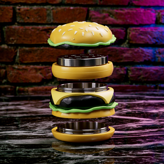 A novelty burger-shaped object shown disassembled into stacked layers, with a yellow bun top and bottom, metallic inner rings, a dark patty layer, and green lettuce-like accents, set on a dark reflective surface with a blurred brick wall background.