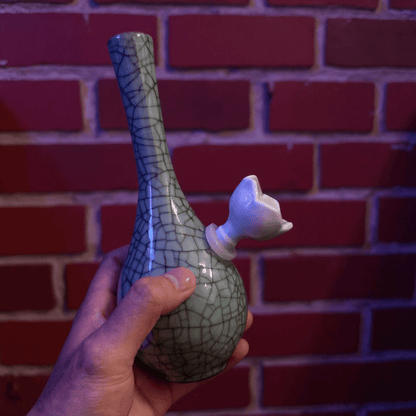 Crackle Glaze Slim Neck Celadon Porcelain BongHand holding a ceramic bong with a cracked glaze design against a brick wall.
