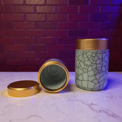 Crackled Blue - Green Celadon Porcelain Stash JarTwo celadon porcelain containers with gold lids on a marble surface against a brick wall.