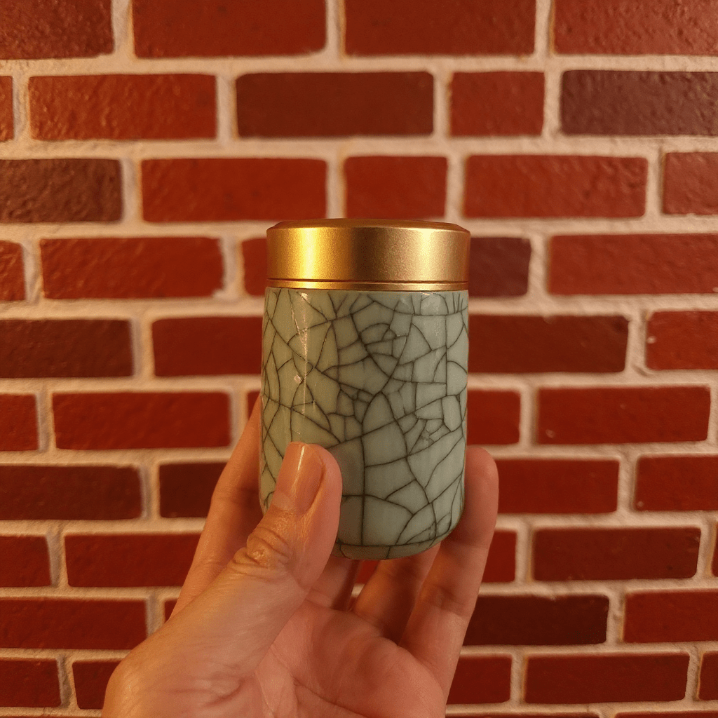 Crackled Blue - Green Celadon Porcelain Stash JarHand holding a crackled celadon porcelain stash jar container with a gold lid against a brick wall.
