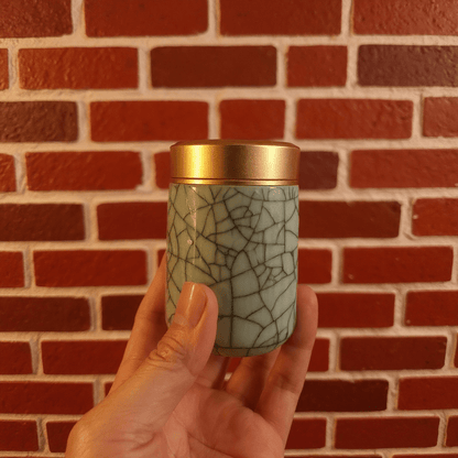 Crackled Blue - Green Celadon Porcelain Stash JarHand holding a crackled celadon porcelain stash jar container with a gold lid against a brick wall.