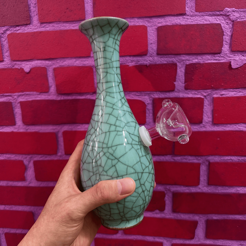 Crackled Light Green Celadon Porcelain Bong – Elegant Ceramic BongGreen ceramic pipe with a clear glass bowl against a red brick wall.