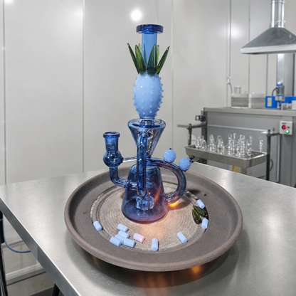 A blue glass water pipe with a pineapple-shaped top section and green leaf details, displayed on a round tray in a clean laboratory or workshop setting.