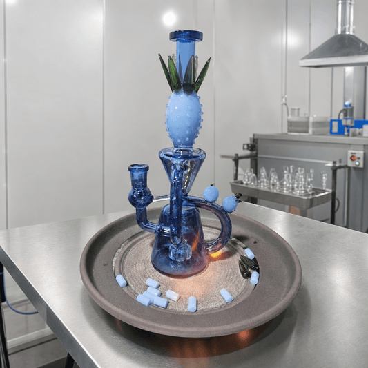 Custom Glass Bong – Personalized Handmade Glass BongA blue glass water pipe with a pineapple-shaped top section and green leaf details, displayed on a round tray in a clean laboratory or workshop setting.