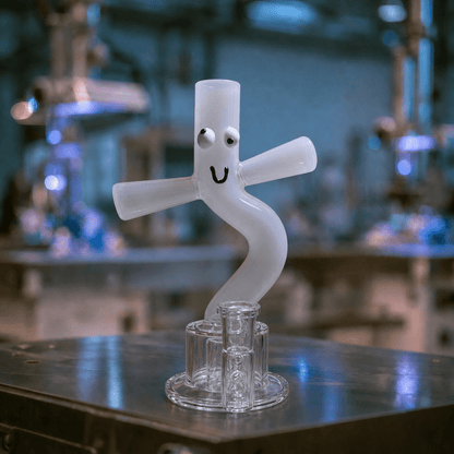 Custom Glass Bong – Personalized Handmade Glass BongA sculptural frosted glass object with a curved, snake-like form and cartoon eyes/mouth, displayed on a clear glass stand on a metal workbench with a blurred laboratory background.