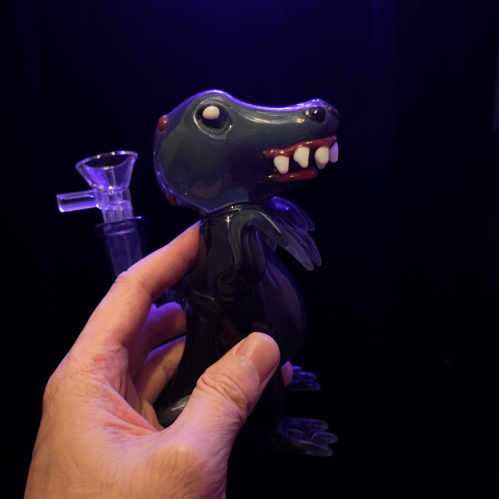 DinoRex Glass Bong - Borosilicate Glass DragonHand holding a glass smoking bong shaped like a dinosaur on a black background