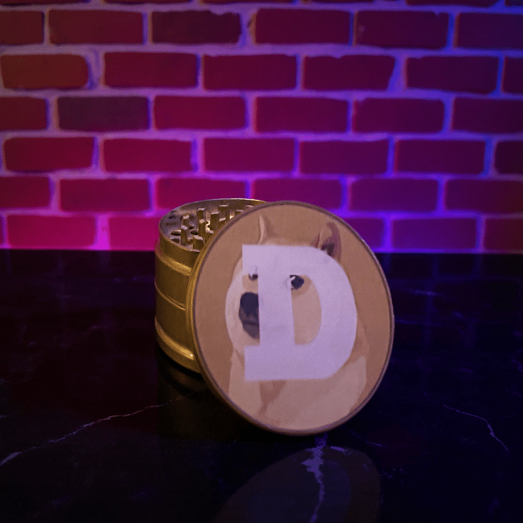 Doge Meme Herb Grinder - Gold - colored grinder with a dog design on a dark surface against a brick wall background