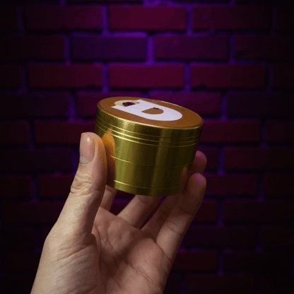 Doge Meme Herb Grinder - Gold - colored grinder with a logo held in a hand against a brick wall background