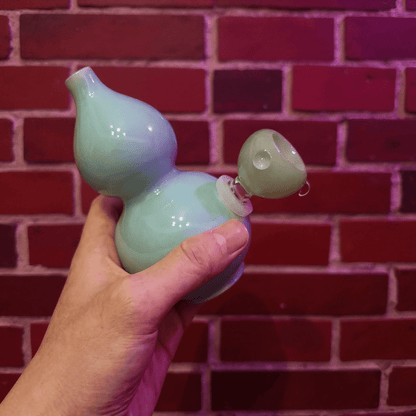 Glossy Blue Celadon Gourd Porcelain BongHand holding a light blue ceramic gourd - shaped bong against a red brick wall.