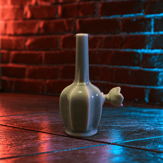 Glossy Green Flat-Sided Celadon Ceramic BongA glossy pale green ceramic bottle or perfume vessel with a tall narrow neck and a small flower-shaped stopper, standing on a dark wooden surface against a red and blue-lit brick wall.