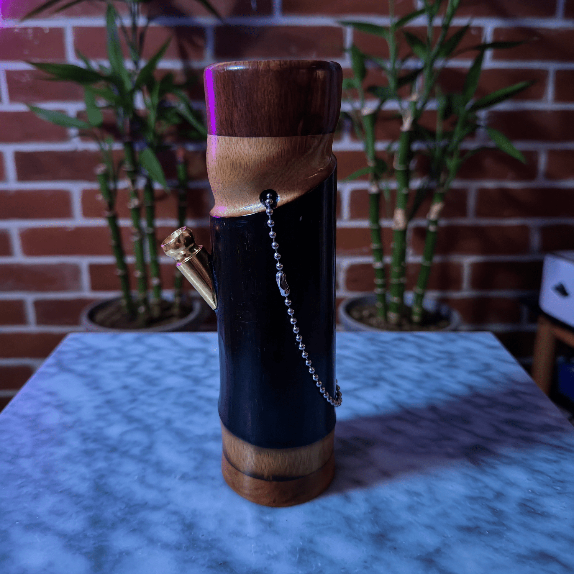 Handcrafted Bamboo Bong – Eco - Friendly Water Pipe with Natural AromaWooden bamboo bong with a chain on a marble surface with plants in the background