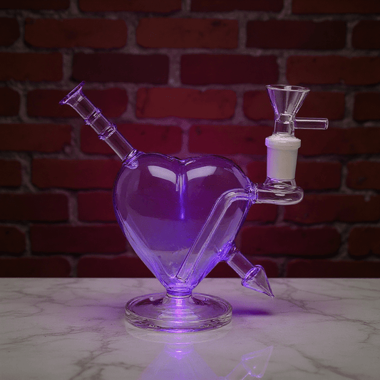 Heartpiercer glass bong - Purple VersionGlass heart - shaped water pipe with a white valve on a marble surface against a brick wall.