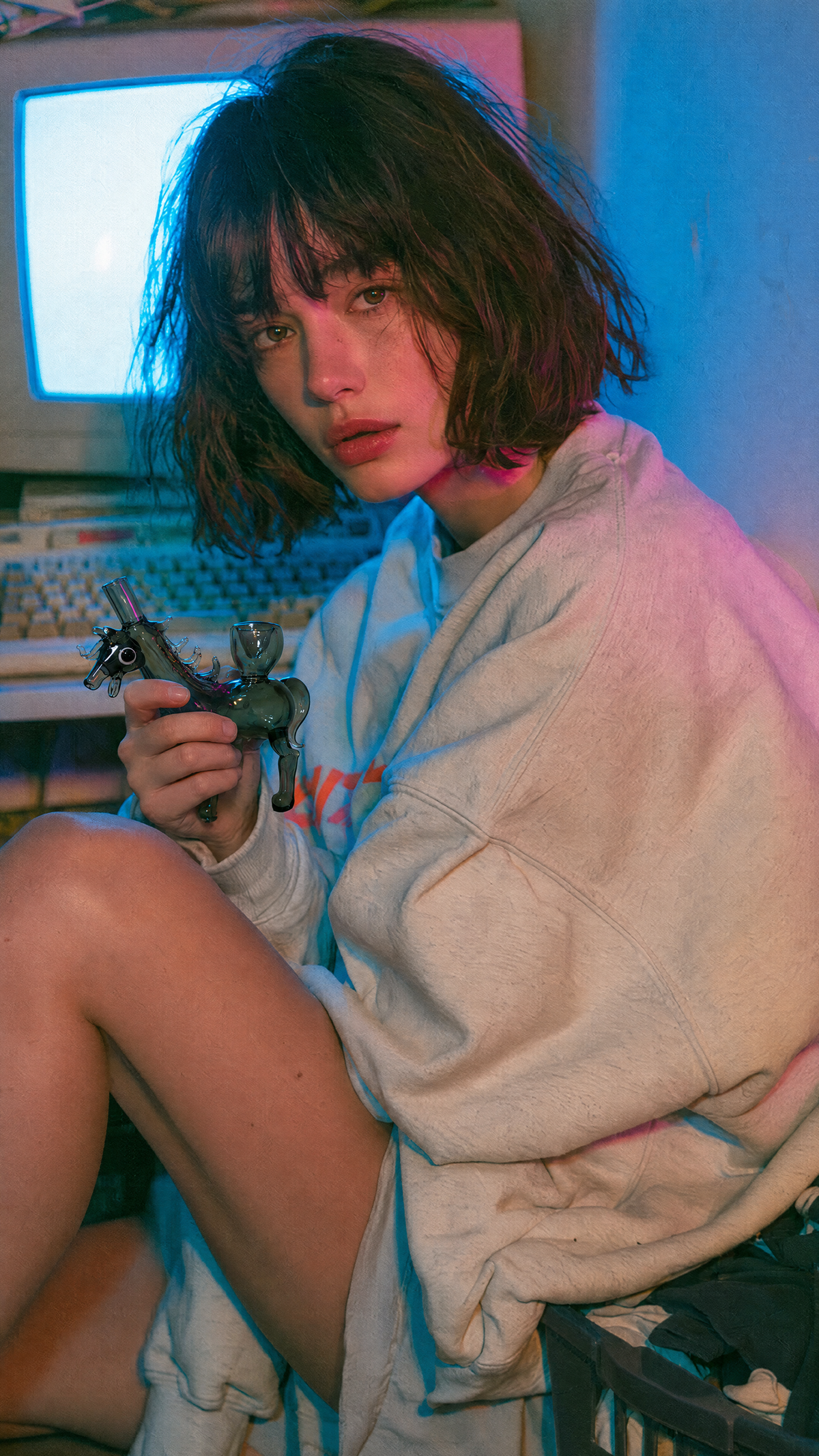 A person with short dark hair sits in a dimly lit room, wrapped in a light gray hoodie and holding a toy gun. A CRT television, keyboard, and desk are visible in the background under blue and pink lighting.