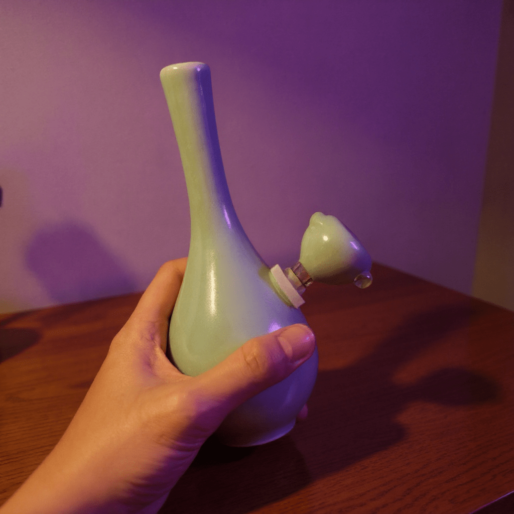 Light Green Slim Neck Celadon Porcelain BongLight green ceramic pipe held by a hand on a wooden surface with a purple background