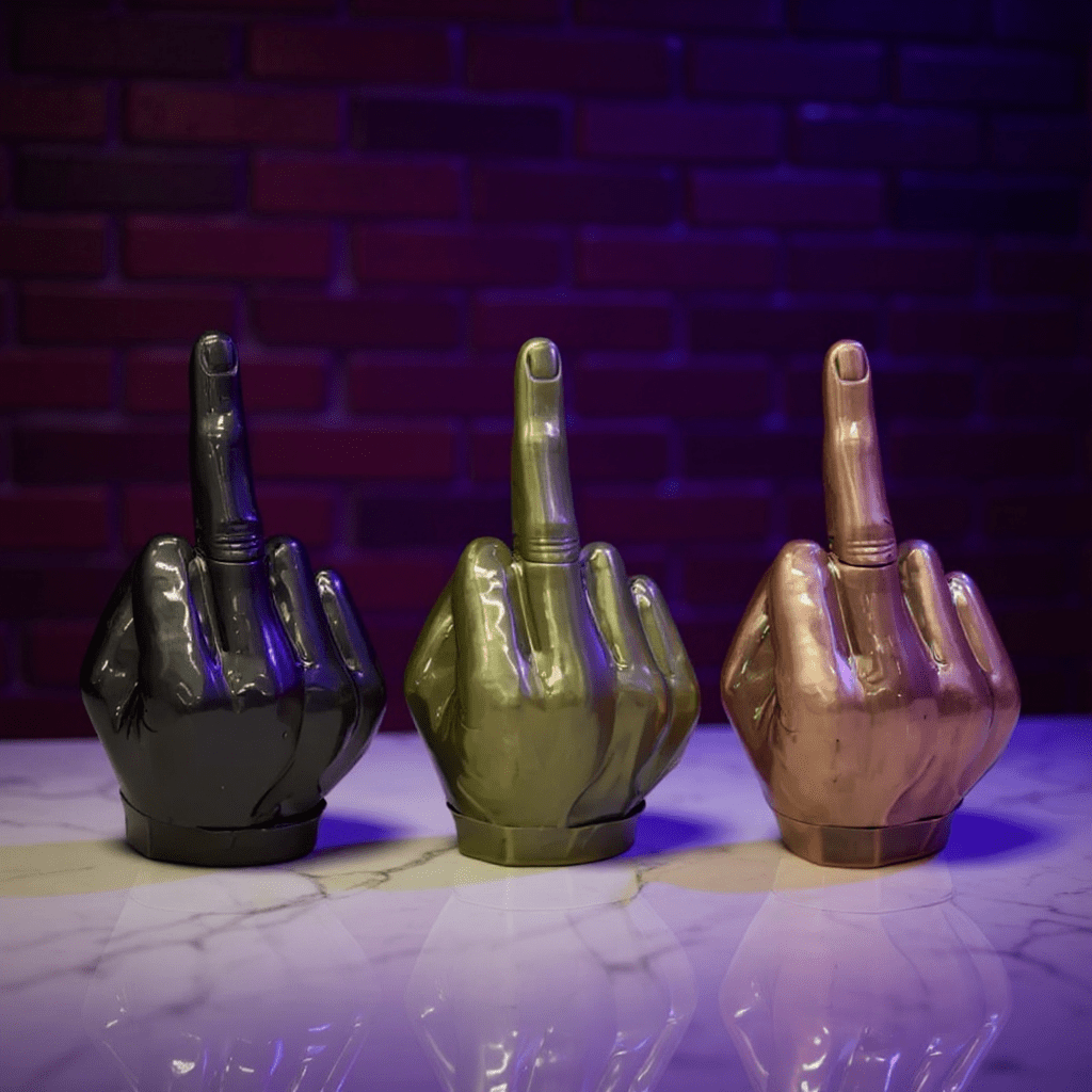 Middle Finger Grinder & Pipe Combo – Heavy Duty 2 - in - 1 SystemThree metallic middle finger grinder in black, green, and bronze on a reflective surface with a brick wall background.