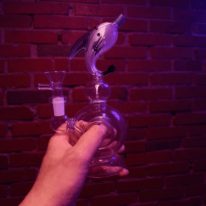 Mythical Creature Glass Bong – Fantasy StyleGlass smoking bong with dolphin design held by a hand against a brick wall.