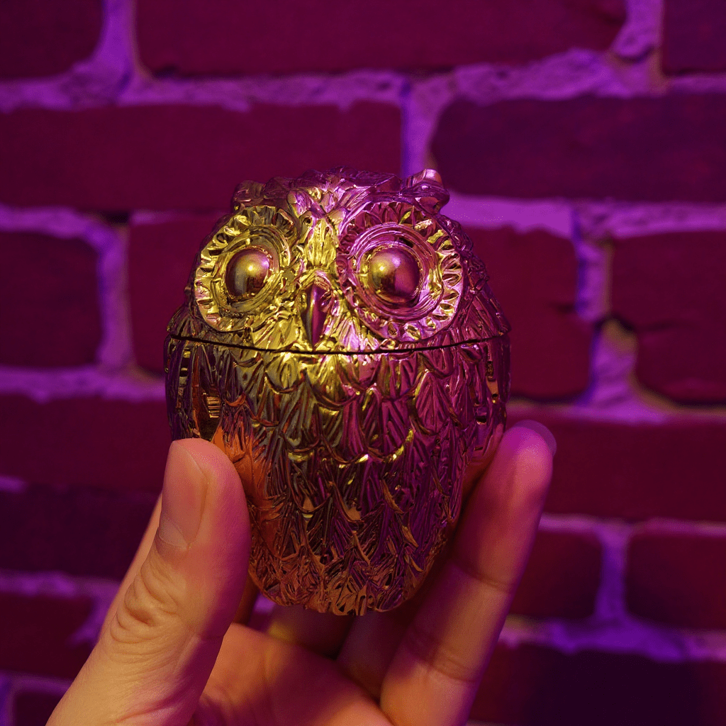 Gold owl - shaped grinder held in front of a brick wall with a purple hue.