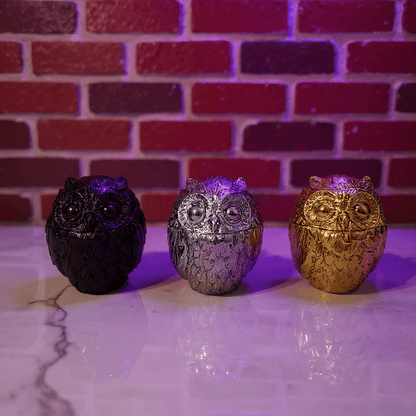 Three owl - shaped grinder in black, silver, and gold against a brick wall background.