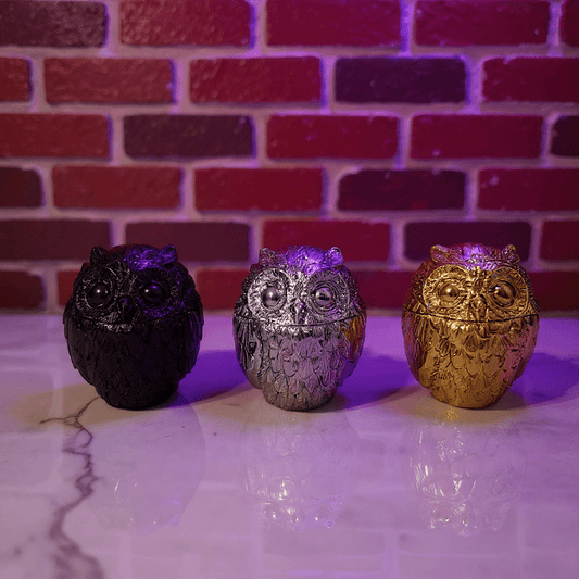 Three owl - shaped grinder in black, silver, and gold against a brick wall background.