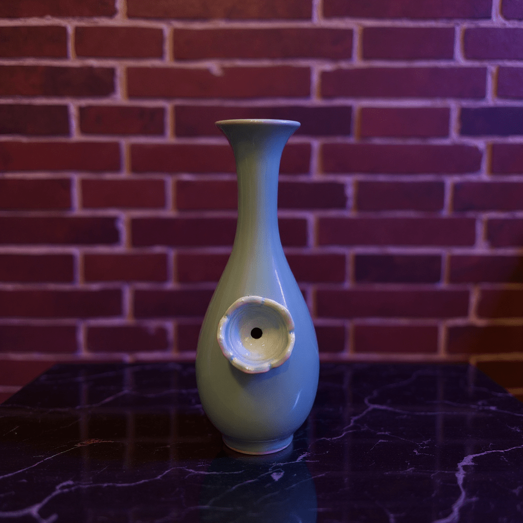 Pale Blue Flared Top Celadon Porcelain BongGreen ceramic bong with a decorative element on a dark surface against a brick wall.