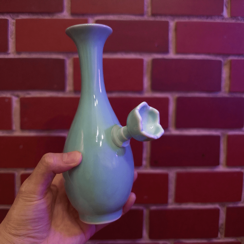 Pale Blue Flared Top Celadon Porcelain BongLight blue ceramic bong with a unique design held against a red brick wall.