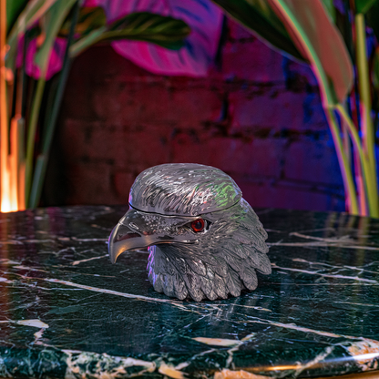 Patriot Eagle Head Metal Grinder – Brass, Bronze & Black FinishA metallic silver eagle head container or sculpture with a textured feathered surface and a small red eye, displayed on a dark green marble surface with a blurred brick wall and plants in the background.