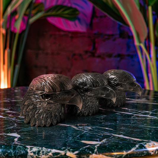 Patriot Eagle Head Metal Grinder – Brass, Bronze & Black FinishA hand holding a bamboo cylindrical water pipe with dark spots, a small brass mouthpiece, and a chain attached, set against a dark brick wall with pink lighting and plants.