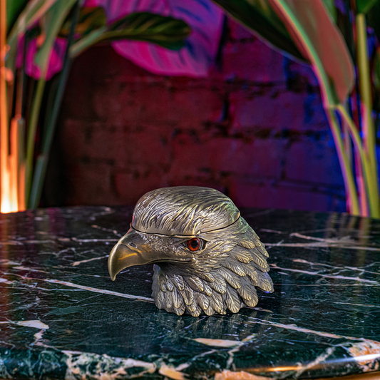 A metallic eagle head container or box with a textured silver finish and a small red eye detail, resting on a dark green marble surface with a blurred brick wall and colorful plants in the background.