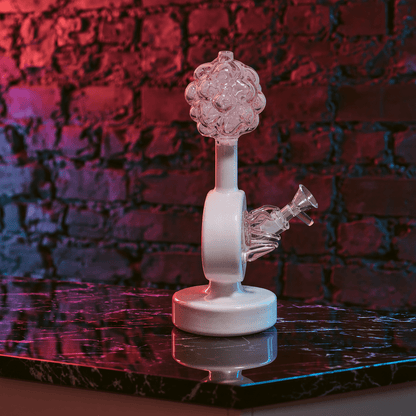 Plumbus-Inspired Sculptural BongA hand holds a decorative cylindrical bamboo box with black branch-like engravings, a gold frog clasp, and a beaded strap, set against a dark brick wall with purple ambient lighting and plants.